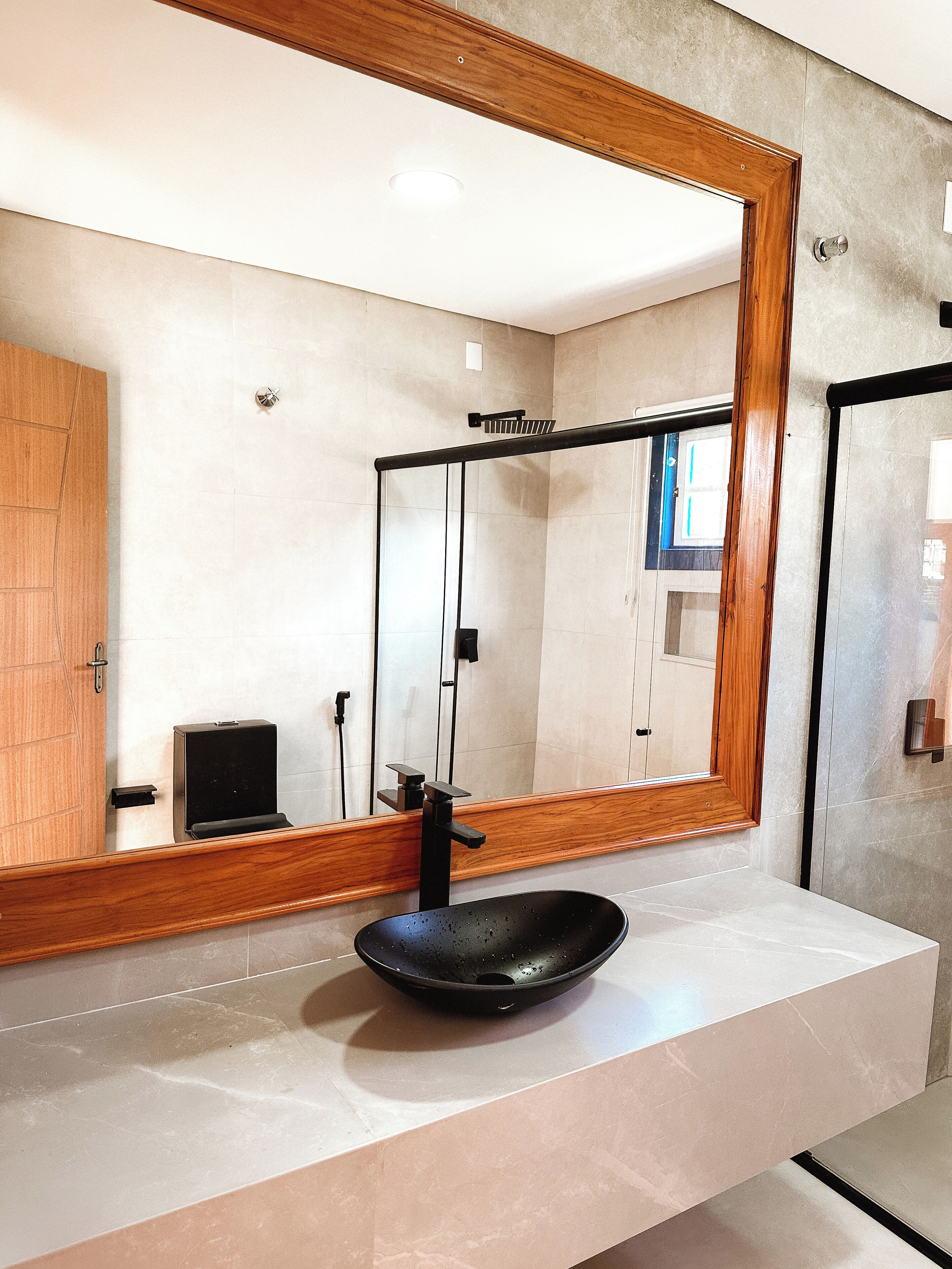 Un baño con un espejo grande, un lavamanos tipo cuenco negro y una puerta de madera.