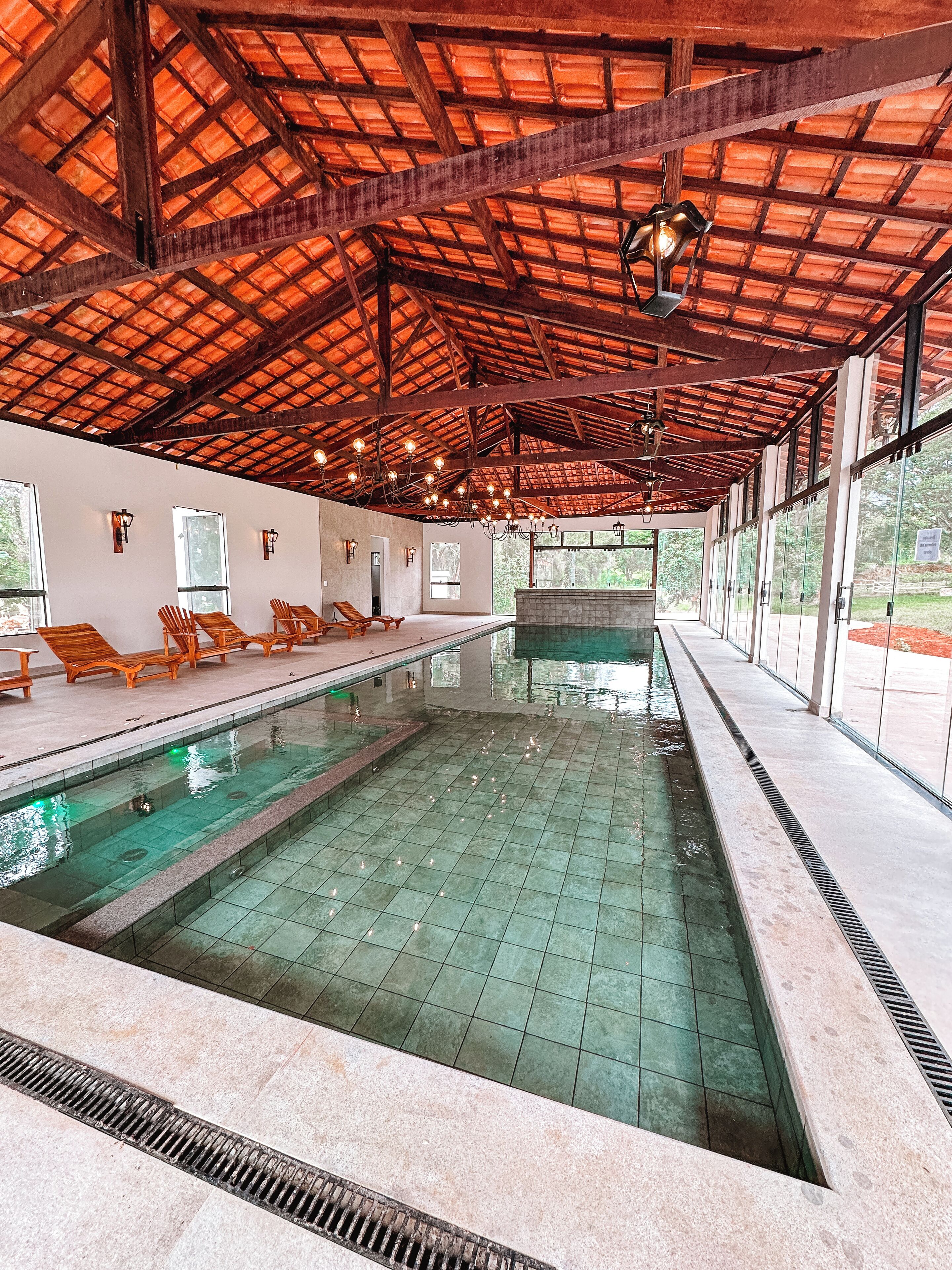 Una piscina interior con suelo de baldosas, sillones de madera y un techo alto con vigas a la vista.