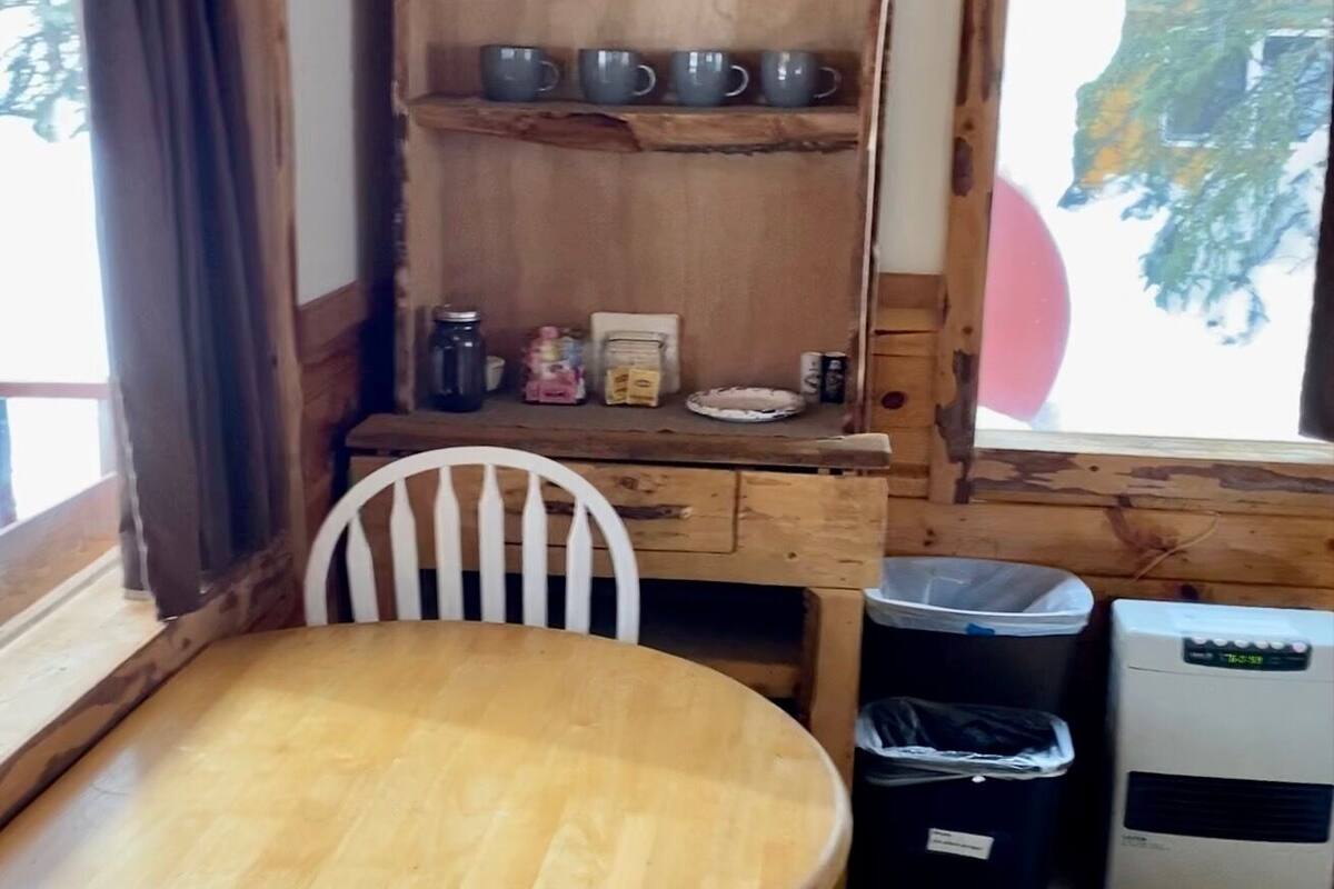 A wooden dining table with a white chair, a shelf with mugs, a trash bin, and a heater.
