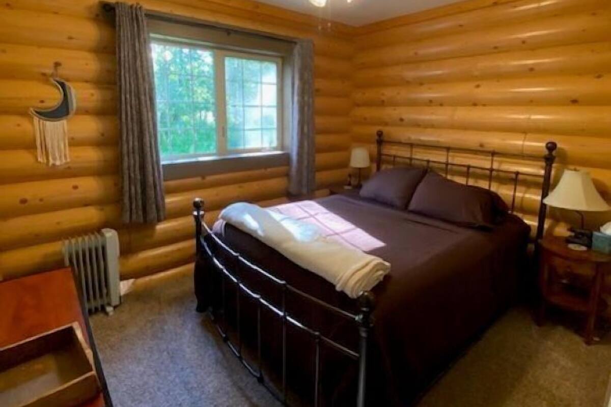 A log cabin bedroom with a single bed, a ceiling fan, a window with curtains, and a small table with a chair.