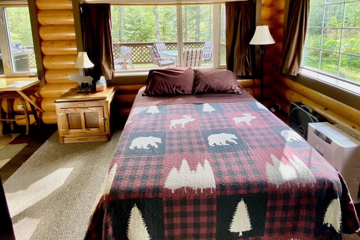 A log cabin bedroom with a bed, a nightstand, a desk, and a window with a view of the outdoors.