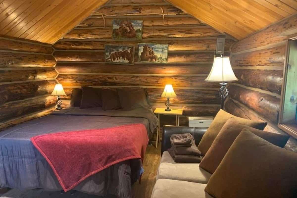 A log cabin bedroom with a bed, two lamps, a desk, and a sofa.