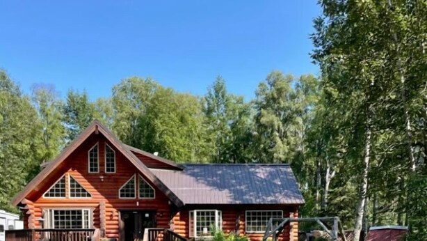 Une cabane en rondins avec un porche, entourée d’une cour verdoyante et d’arbres.