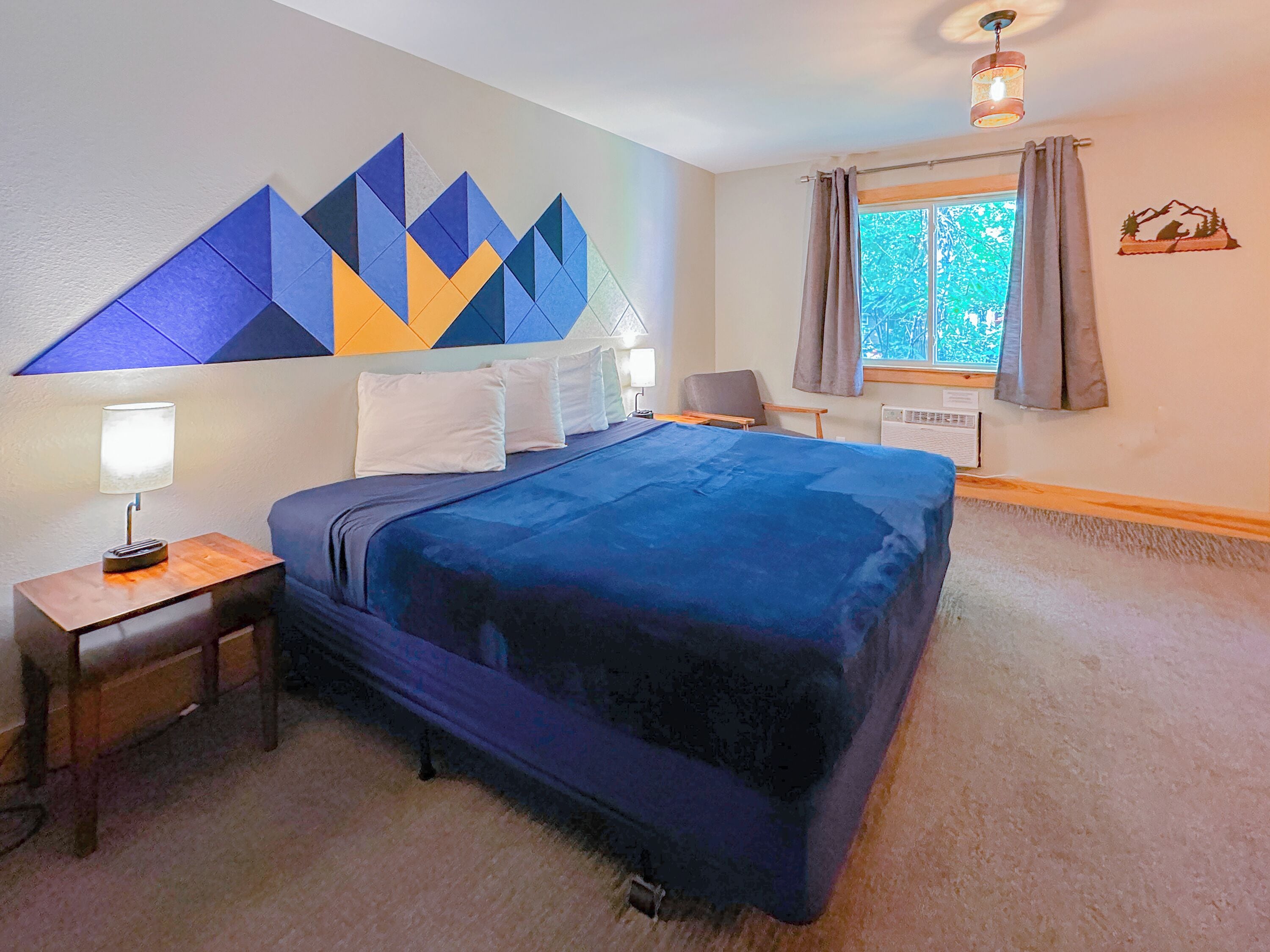 A hotel room with a large bed, a nightstand with a lamp, a window with curtains, and a wall decoration.