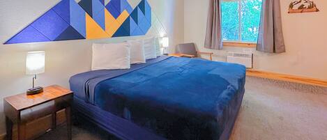 A hotel room with a large bed, a nightstand with a lamp, a window with curtains, and a wall decoration.