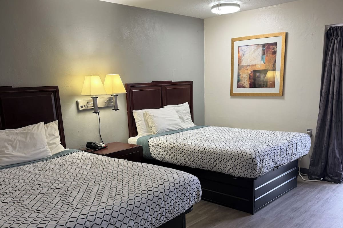 Two beds with patterned bedding, a nightstand with a lamp, a painting on the wall, and a curtain.