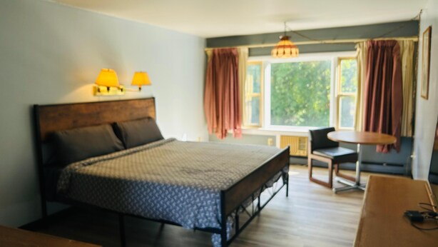A bedroom with a bed, two bedside lamps, a window with curtains, a chair, and a small table. - Capital O Swan Inn Comstock Park (Comstock Park)