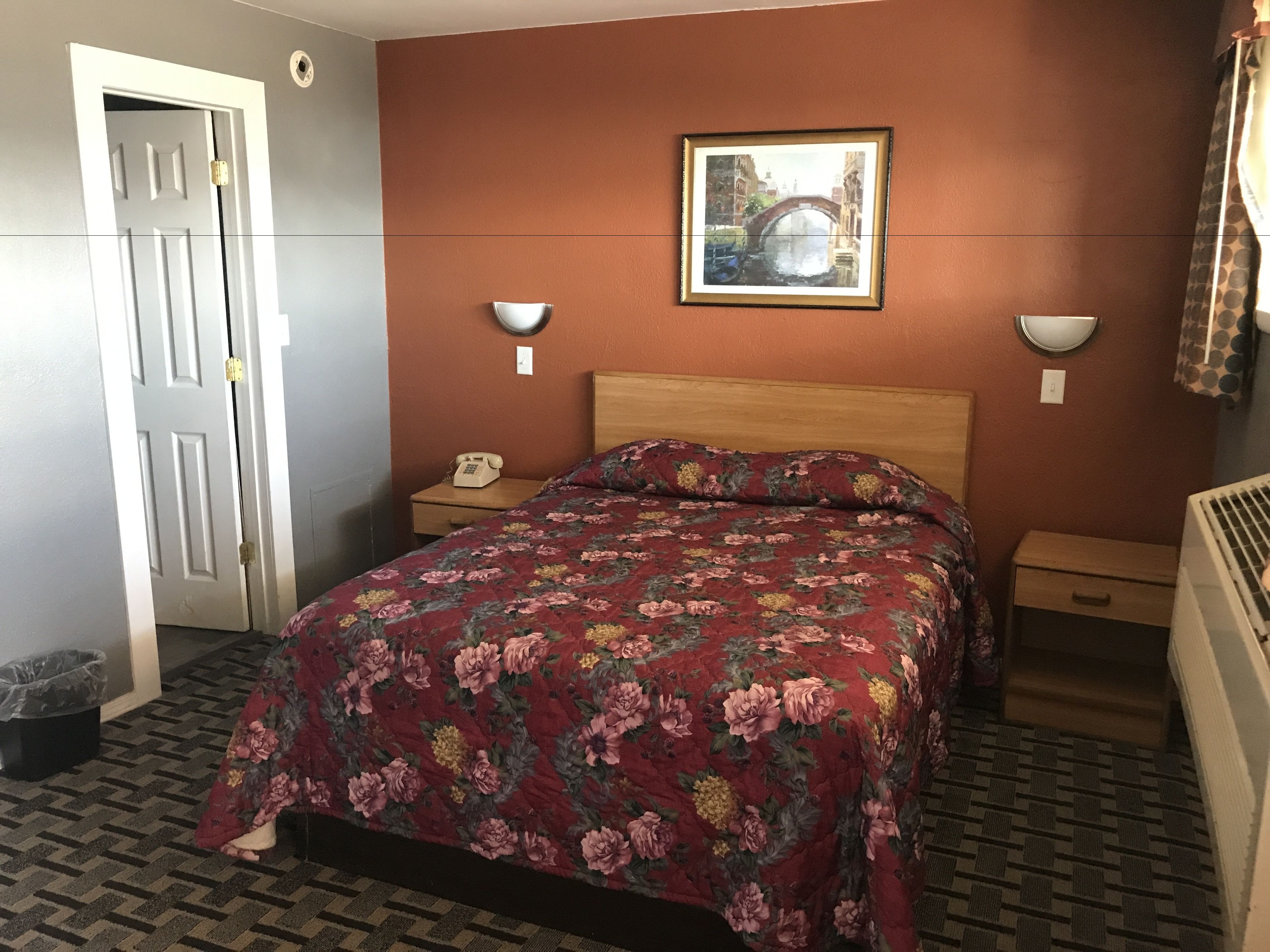 A bedroom with a floral bedspread, a wooden headboard, a bedside table with a lamp, and a framed picture on the wall.