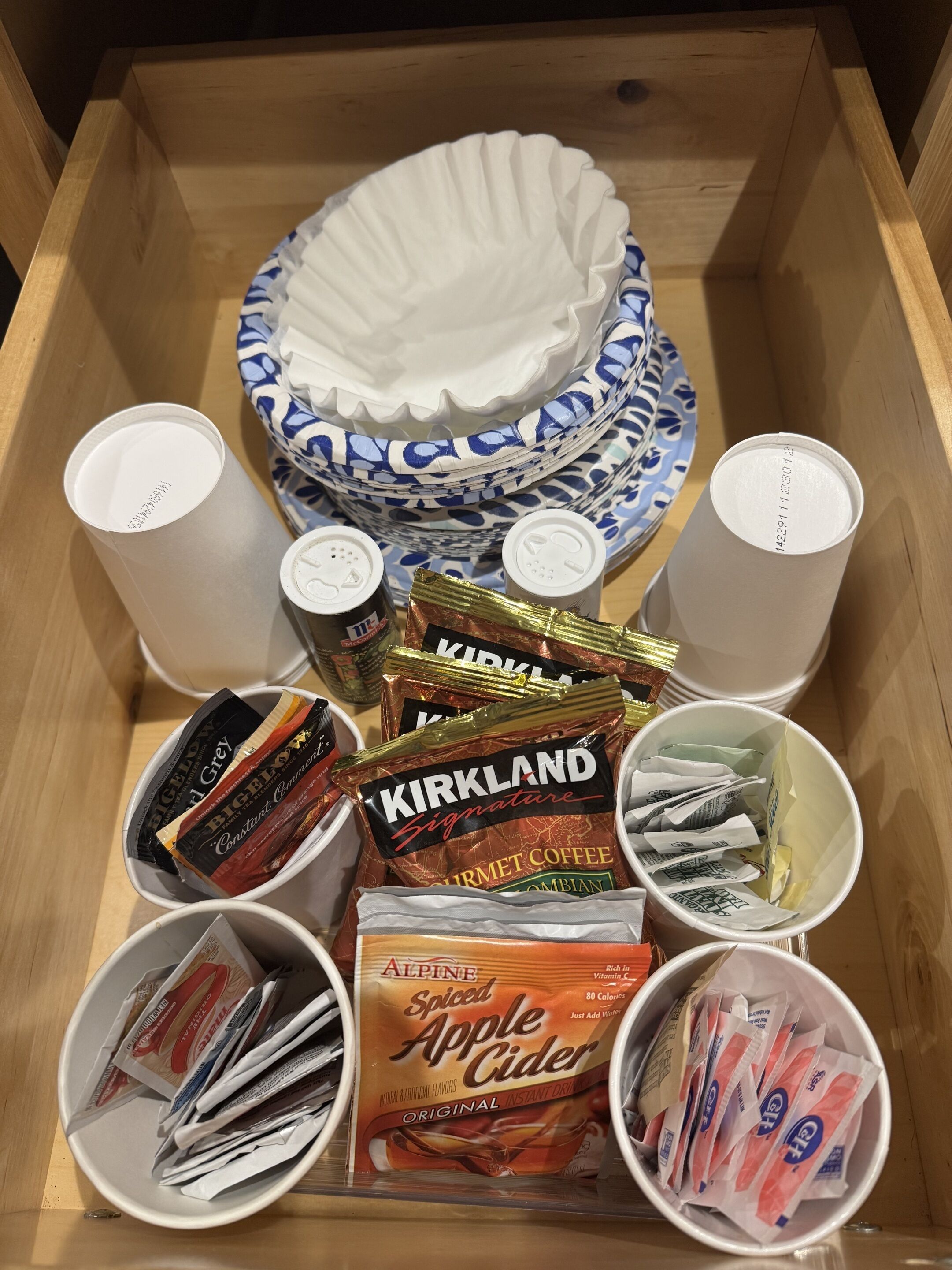 A wooden box containing paper plates, disposable cups, and various food items including Kirkland Signature coffee and Alpine Spiced Apple Cider.