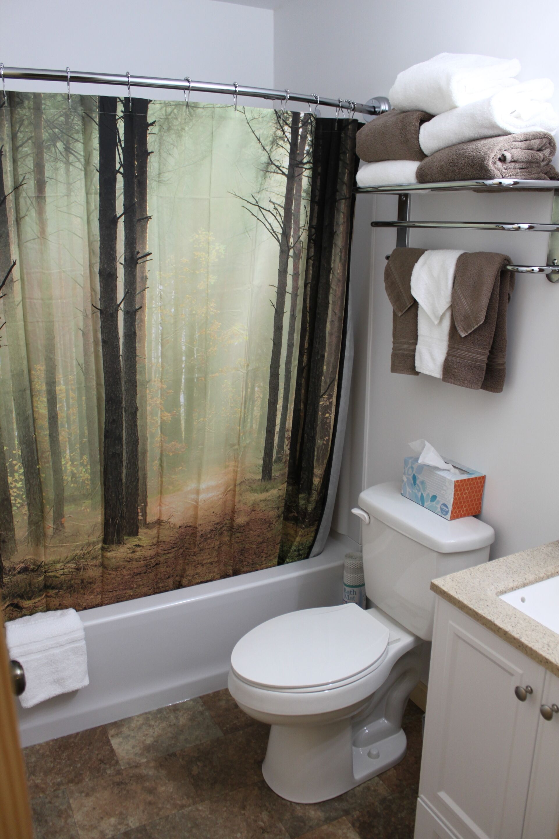 A bathroom with a forest-themed shower curtain, a toilet, a bathtub, and a shelf with towels.