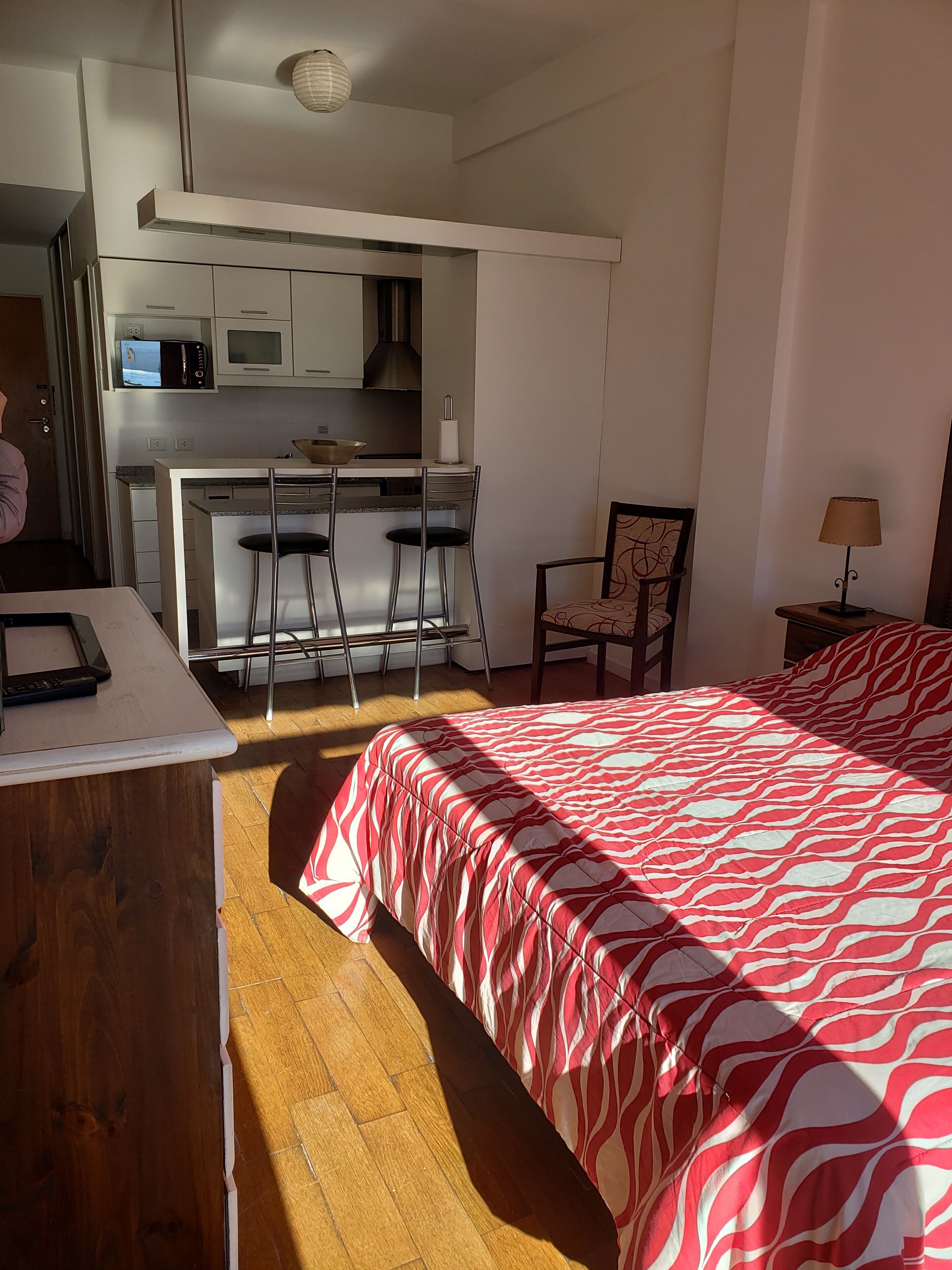 Habitación de hotel compacta con cama, escritorio, silla y kitchenette.