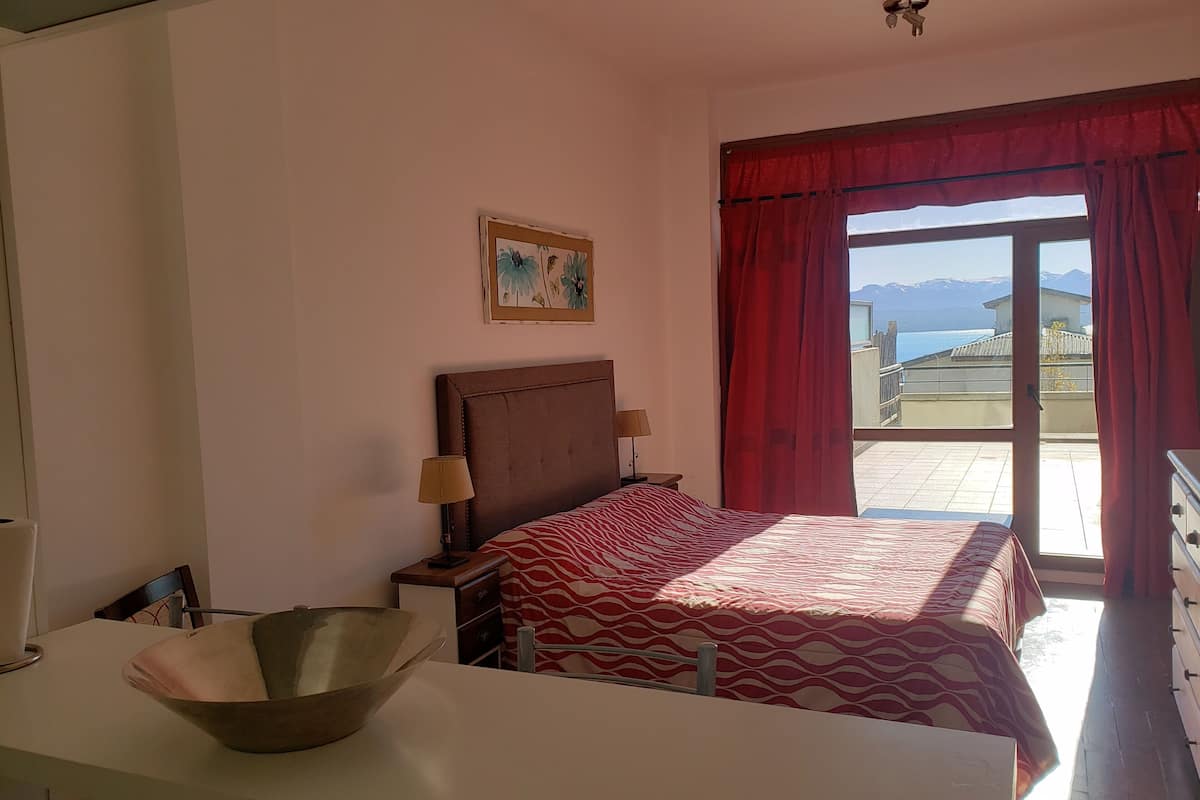 Un dormitorio con cama, mesita de noche, lámpara, un cuenco, un cuadro y vistas al exterior.