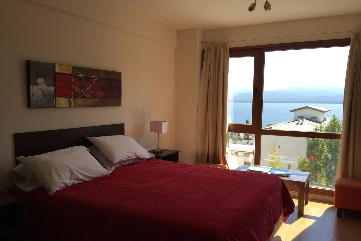 Un dormitorio con una cama grande, una mesita de noche, una lámpara y una ventana con vistas a un cuerpo de agua y edificios.