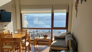 Ett rymligt rum med trägolv, ett matbord med stolar, en soffa, ett litet runt bord med en flaska på och ett stort fönster med utsikt över berg och en sjö eller ett annat vattendrag.