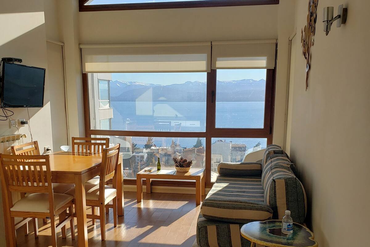Una habitación amplia con suelo de madera, una mesa de comedor con sillas, un sofá, una pequeña mesa redonda con una botella, y un gran ventanal con vistas a montañas y una masa de agua.