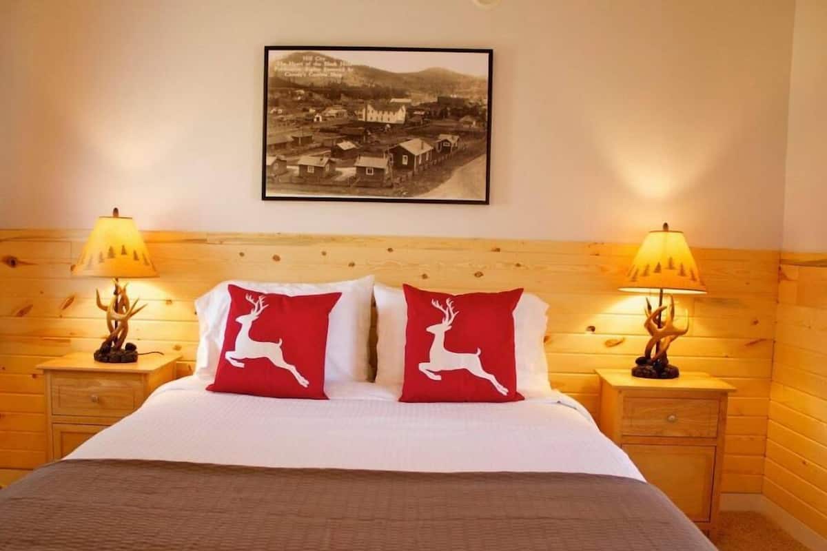 Een hotelkamer met een houten hoofdbord, twee bedlampjes, een bed met wit beddengoed en twee rode kussens, en een ingelijst schilderij aan de muur.