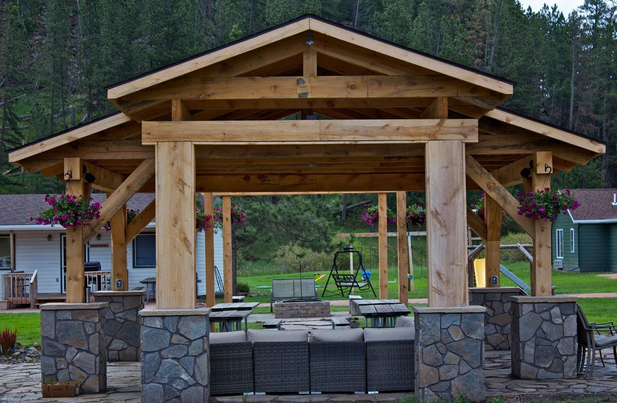 A wooden gazebo with stone pillars and a stone fireplace, surrounded by outdoor seating.