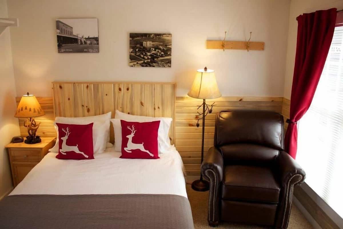 Een hotelkamer met een houten hoofdeinde, twee kussens met hertjes in rood en wit, een bruine leren fauteuil, een nachtkastje met een lamp en twee aan de muur hangende schilderijen.