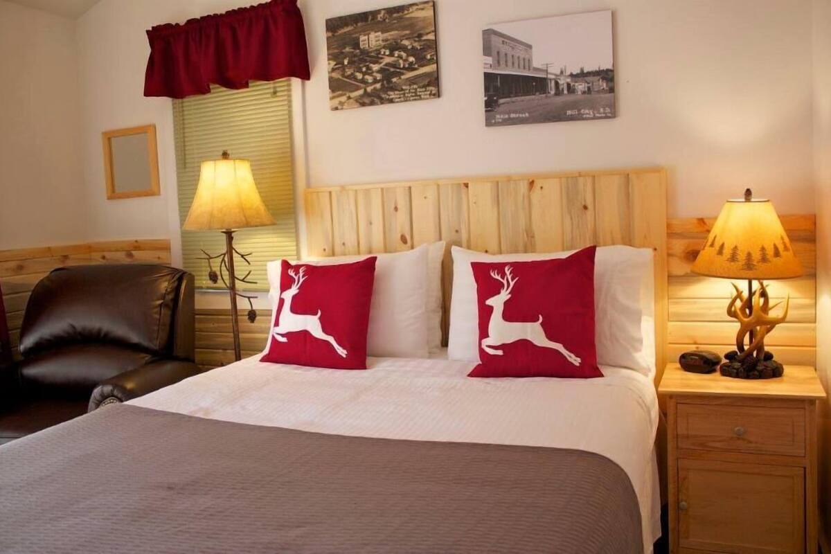 Een hotelkamer met een houten hoofdeinde, twee rode kussens met witte hertenmotieven, een bed met een witte deken, een houten nachtkastje met een lamp en twee ingelijste foto's aan de muur.