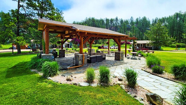 A covered outdoor seating area with stone flooring and wooden furniture, surrounded by a well-maintained lawn and trees. - Black Elk Resort Cottages and RV Park (Hill City)