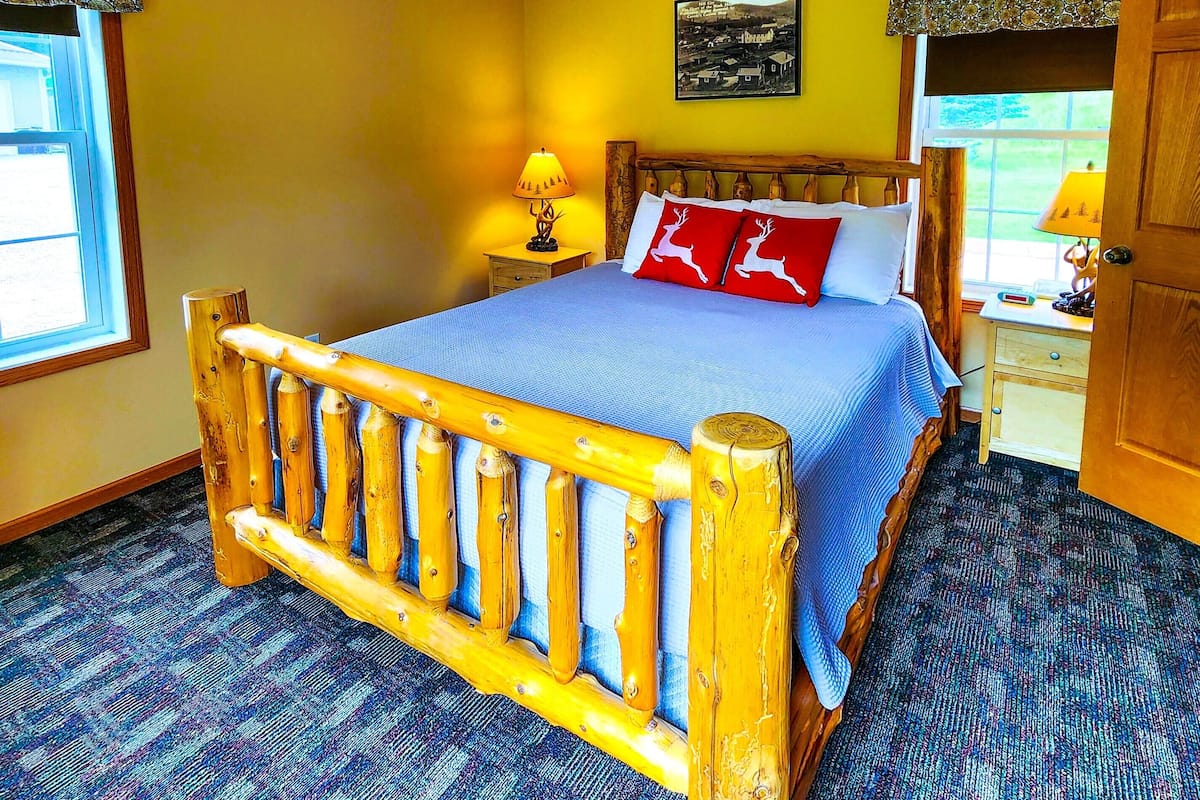 Een bed met houten frame, een blauwe spreideken en twee rode kussens met hertenmotieven. Er staat een nachtkastje met een lamp, een raam met uitzicht en een deur aan de rechterkant.