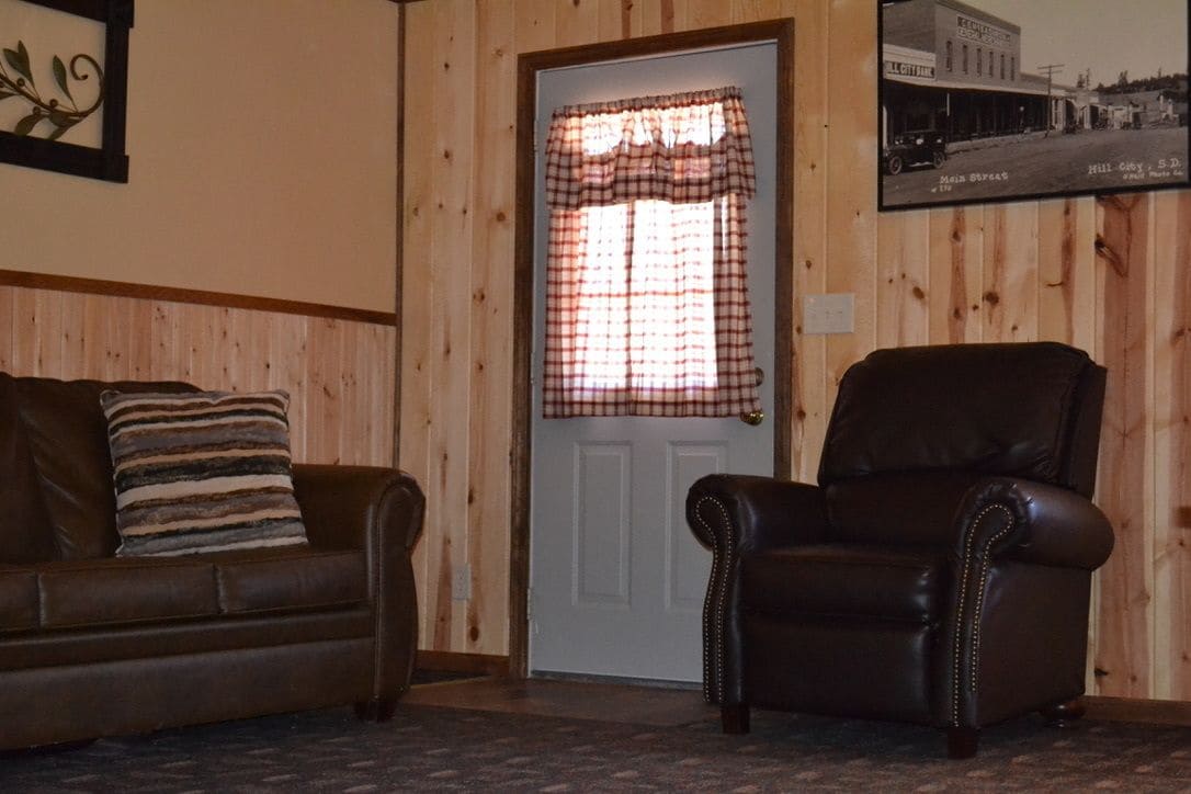 Une pièce avec un mur lambrissé en bois, une porte ornée d’un rideau à carreaux, un canapé en cuir marron avec un coussin rayé et un fauteuil en cuir.