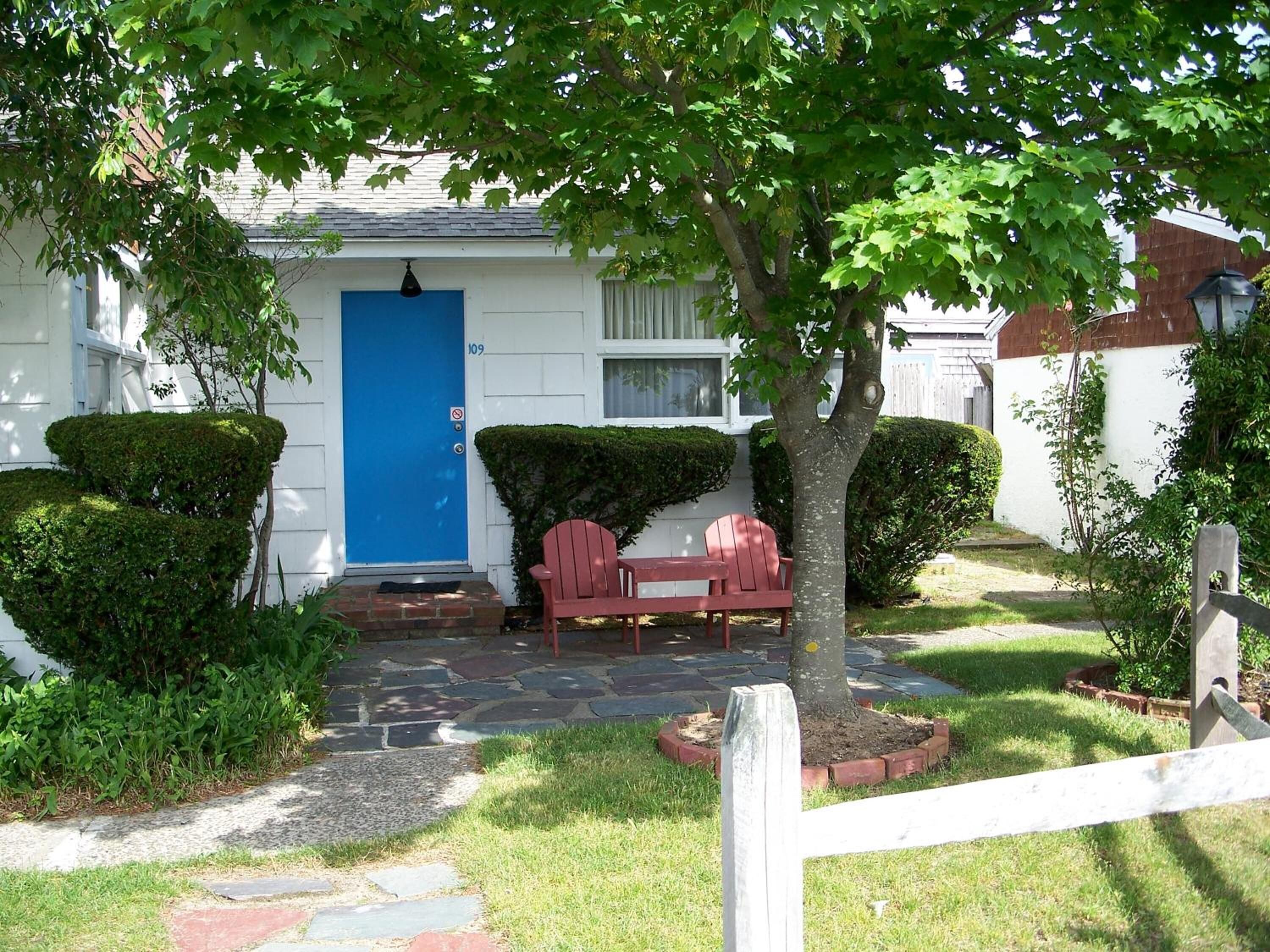 Une maison avec une porte bleue, une chaise rouge et un arbre dans le jardin.