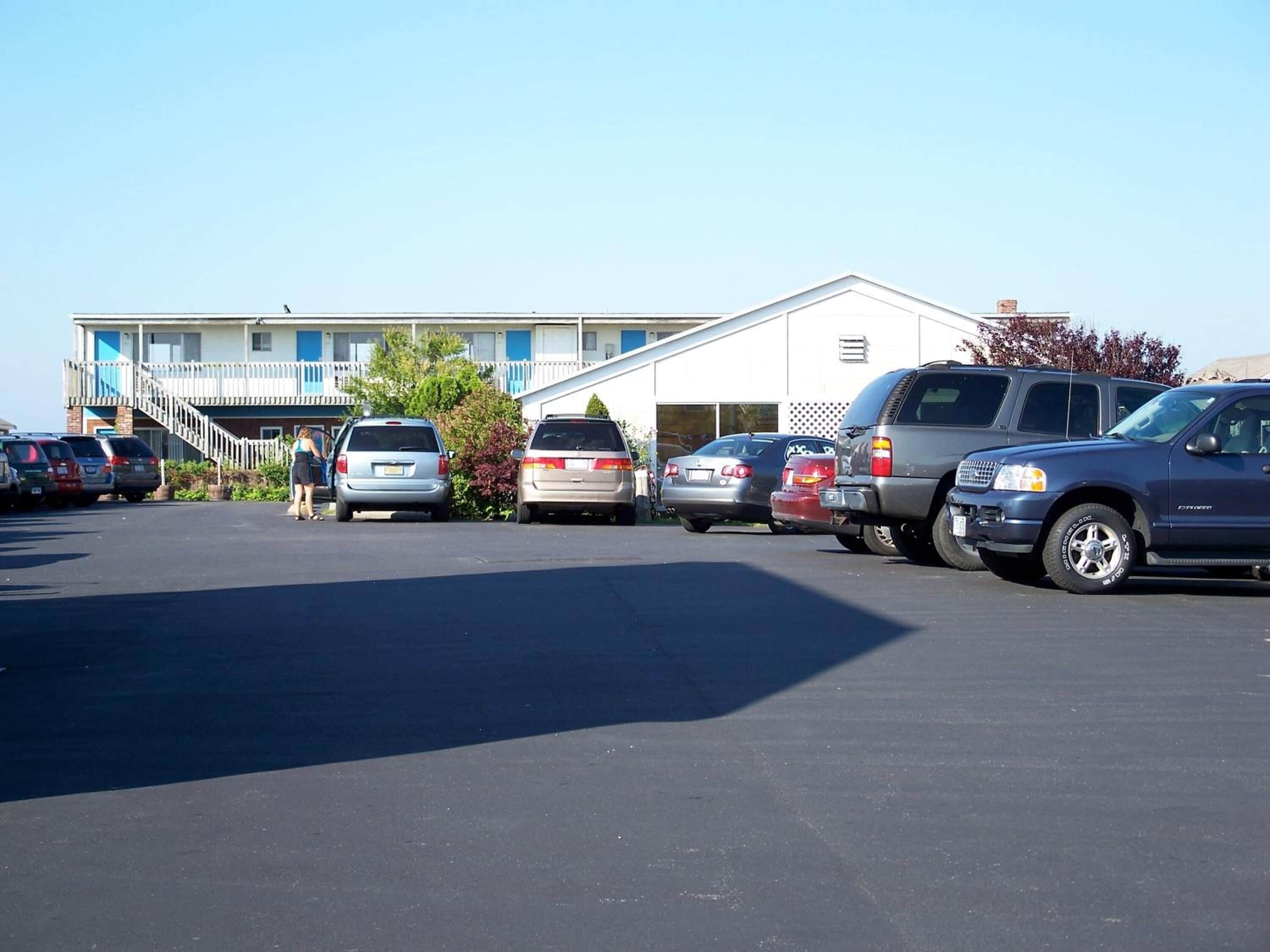 A parking lot with several cars, a building with a balcony, and a person walking.