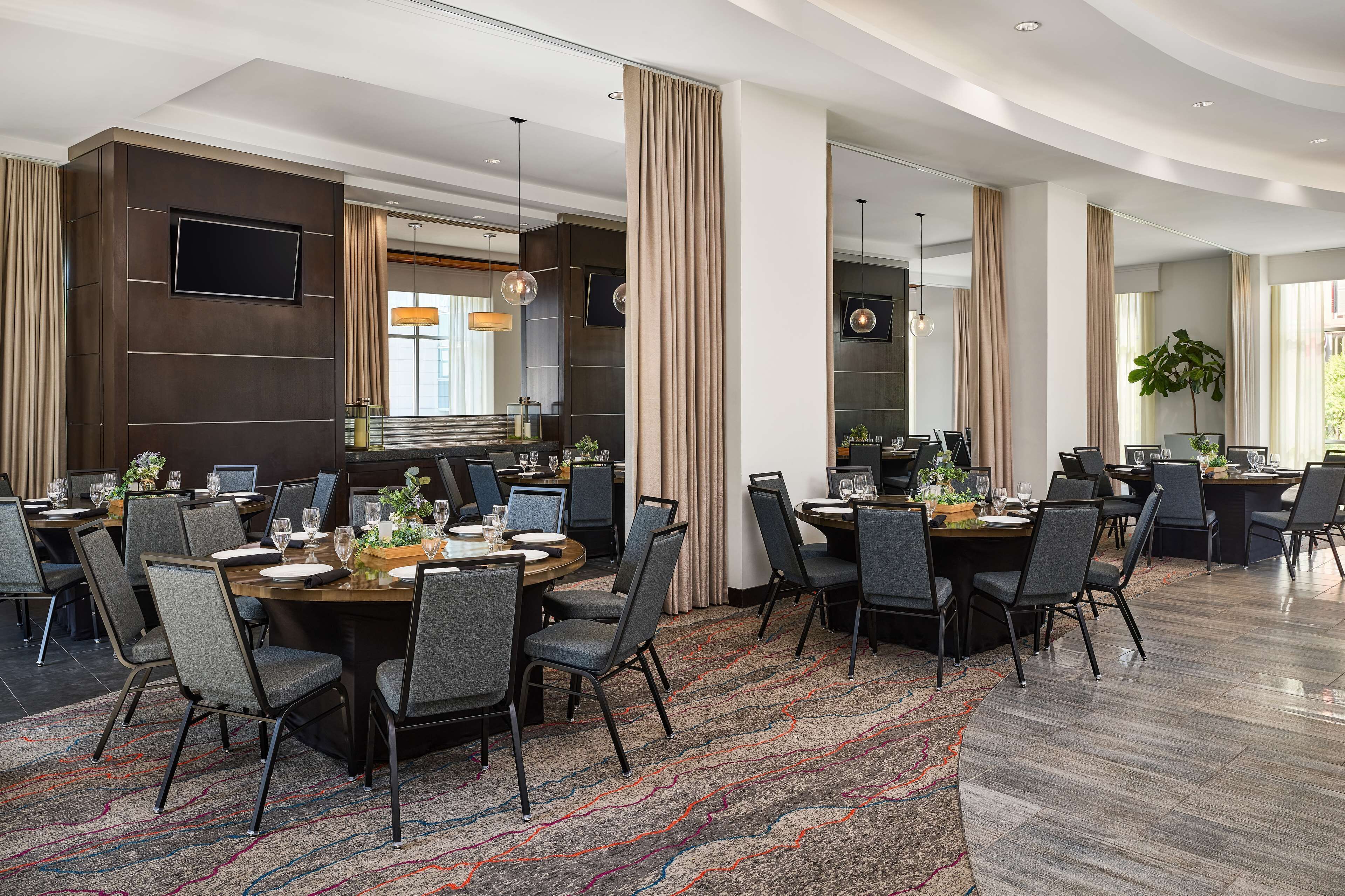 A modern hotel dining area with round tables, grey chairs, and a large mirror.