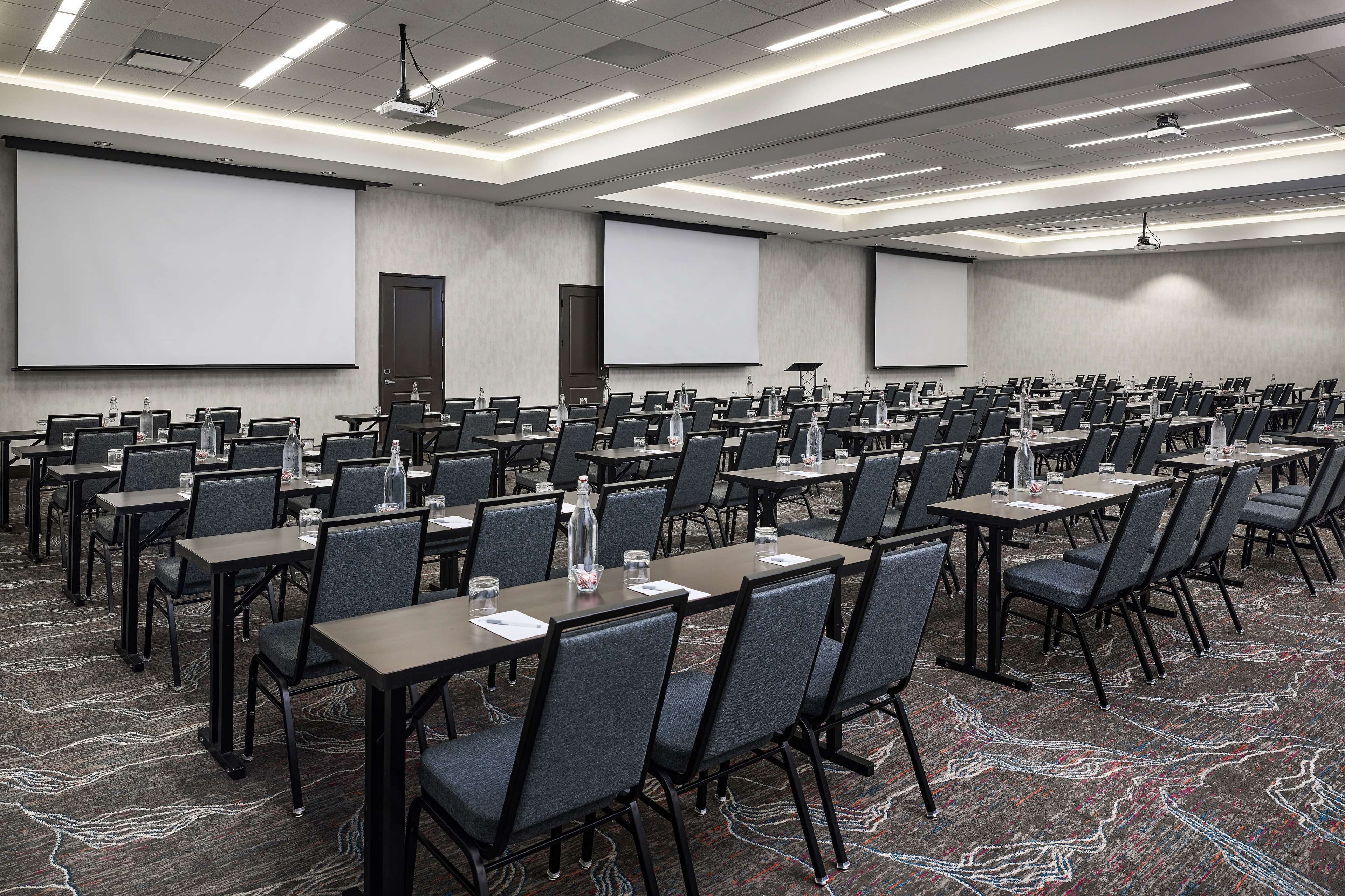 A conference room with rows of tables and chairs, equipped with projectors and screens, and a carpeted floor.