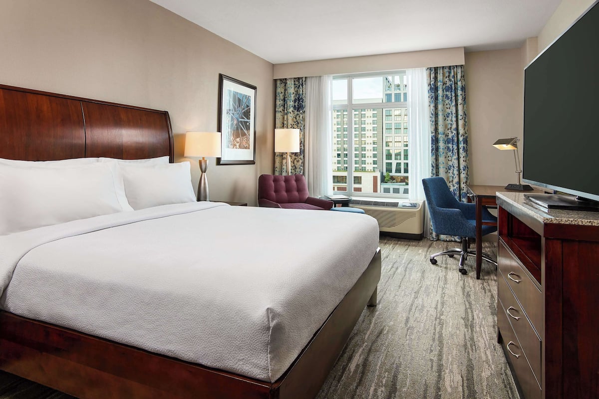 A hotel room with a large bed, a desk with a television, a chair, and a window with curtains.