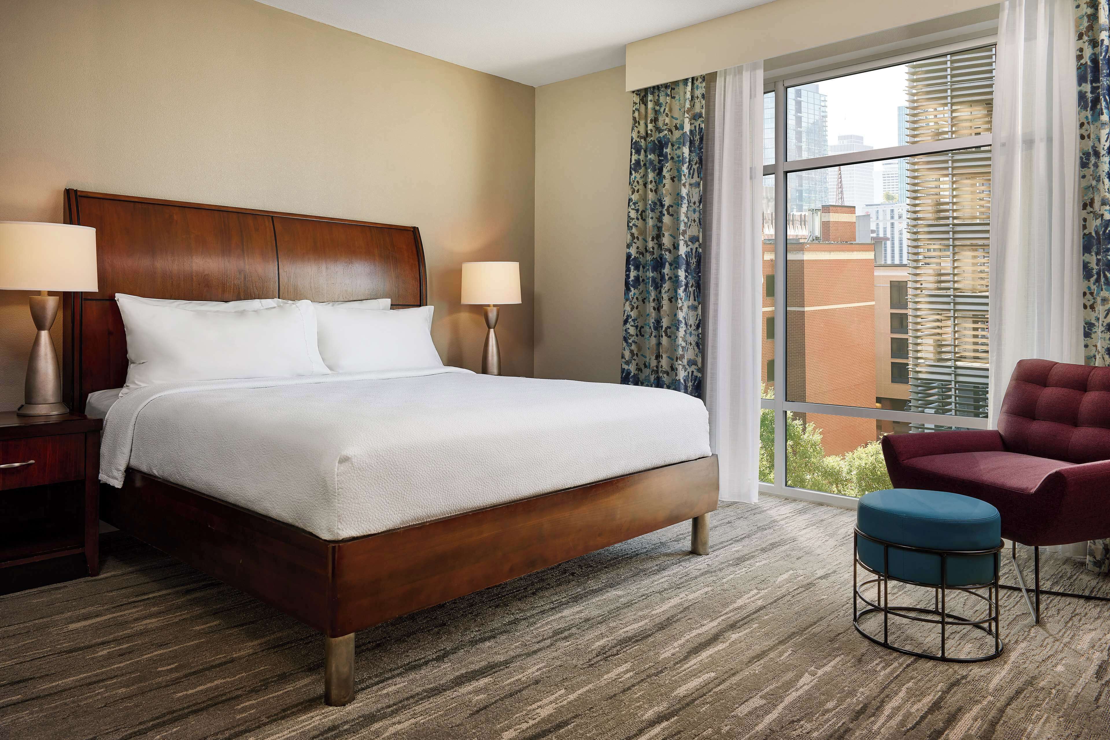 A hotel room with a large bed, two bedside lamps, a window with blinds, a red chair, and a blue ottoman.