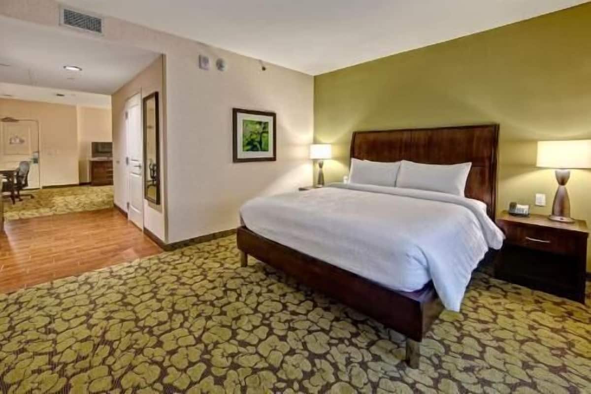 A hotel room with a bed, a nightstand, a lamp, a picture on the wall, and a bathroom visible through an open door.