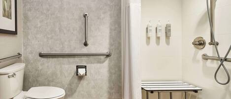 A bathroom with a toilet, a shower with a handheld showerhead, grab bars, and a small table.