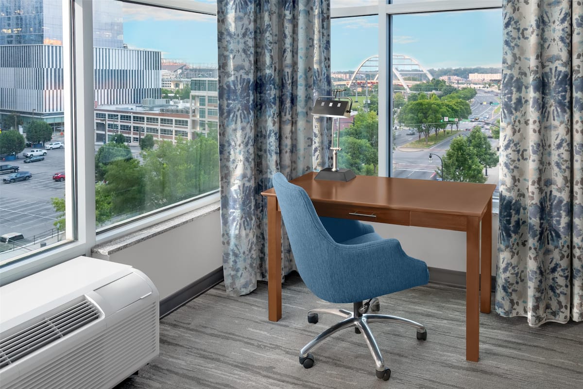 A modern office room with a large window, a desk, a blue office chair, and a white air conditioner.