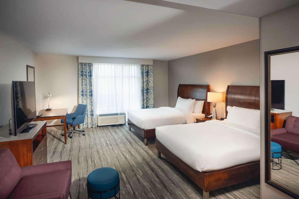 A hotel room with two beds, a desk with a chair, a TV, and a large window with curtains.
