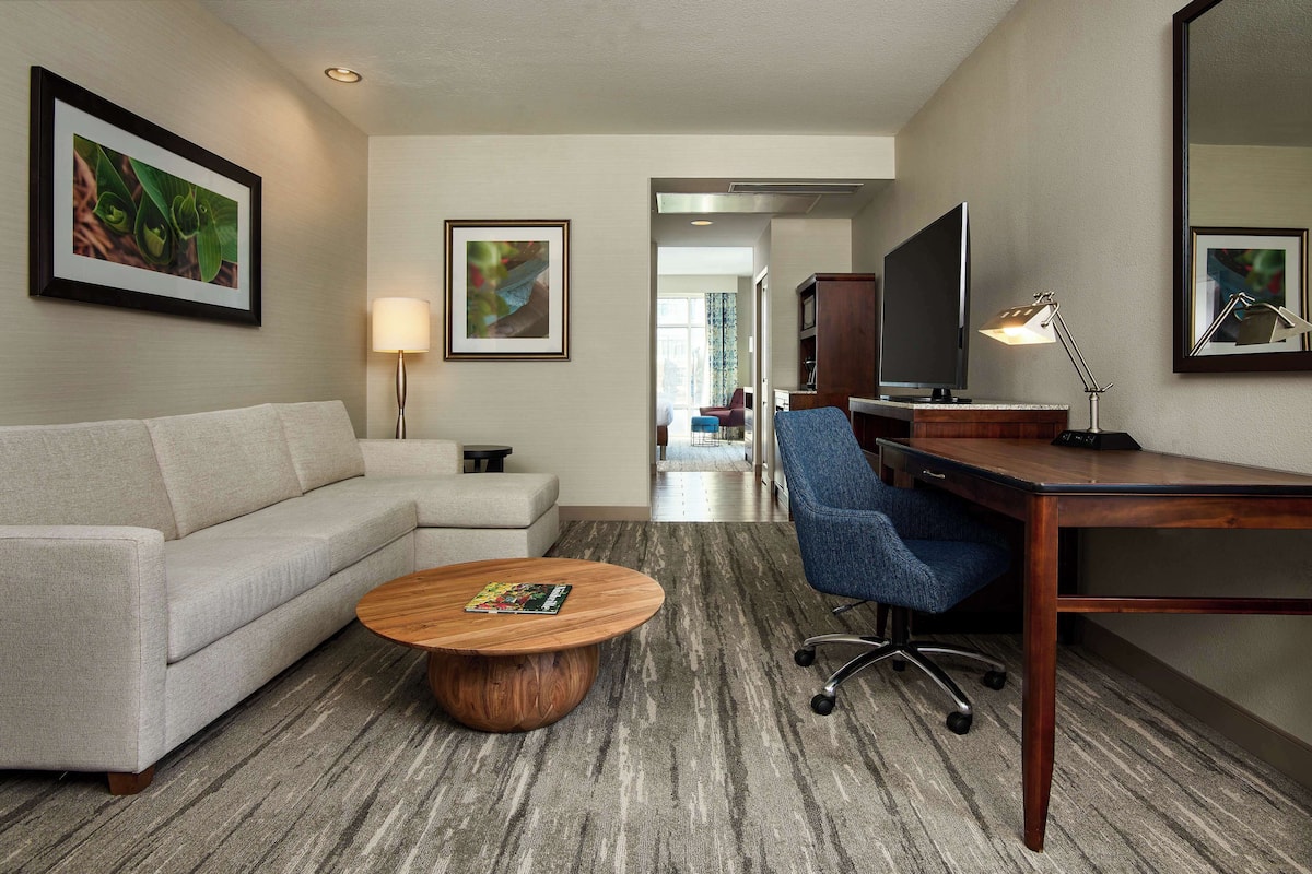 A hotel room with a sofa, a coffee table, a desk with a chair, a TV, and framed artwork on the walls.