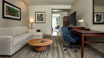 A hotel room with a sofa, a coffee table, a desk with a chair, a TV, and framed artwork on the walls.