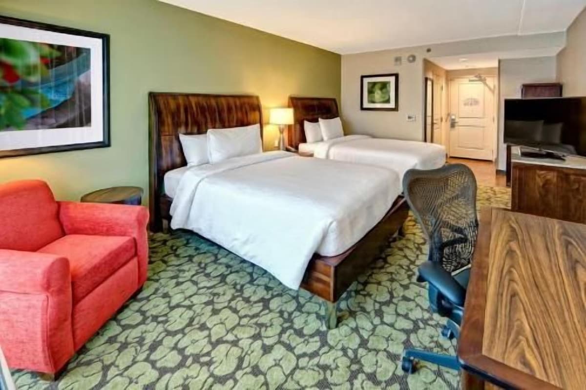 A hotel room with two beds, a red armchair, a wooden desk, and a television.