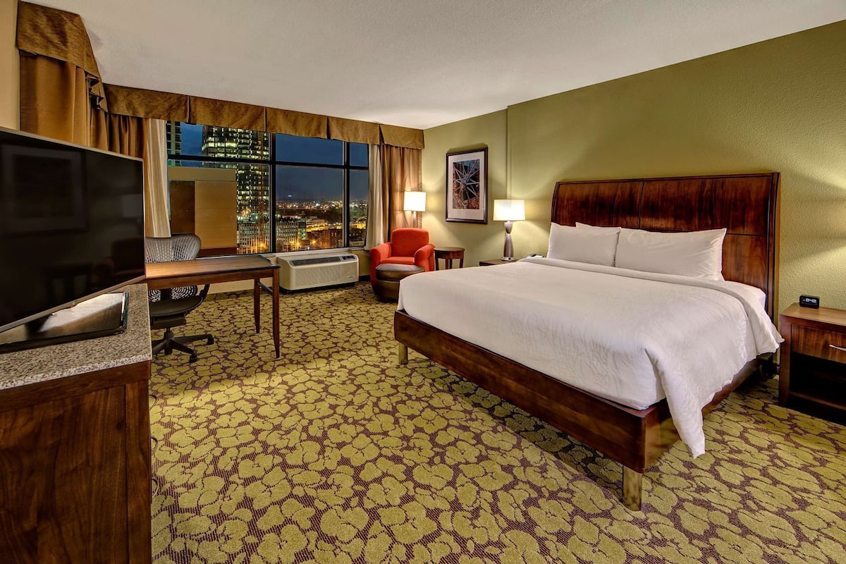 A hotel room with a large bed, a desk, a television, and a view of the cityscape.