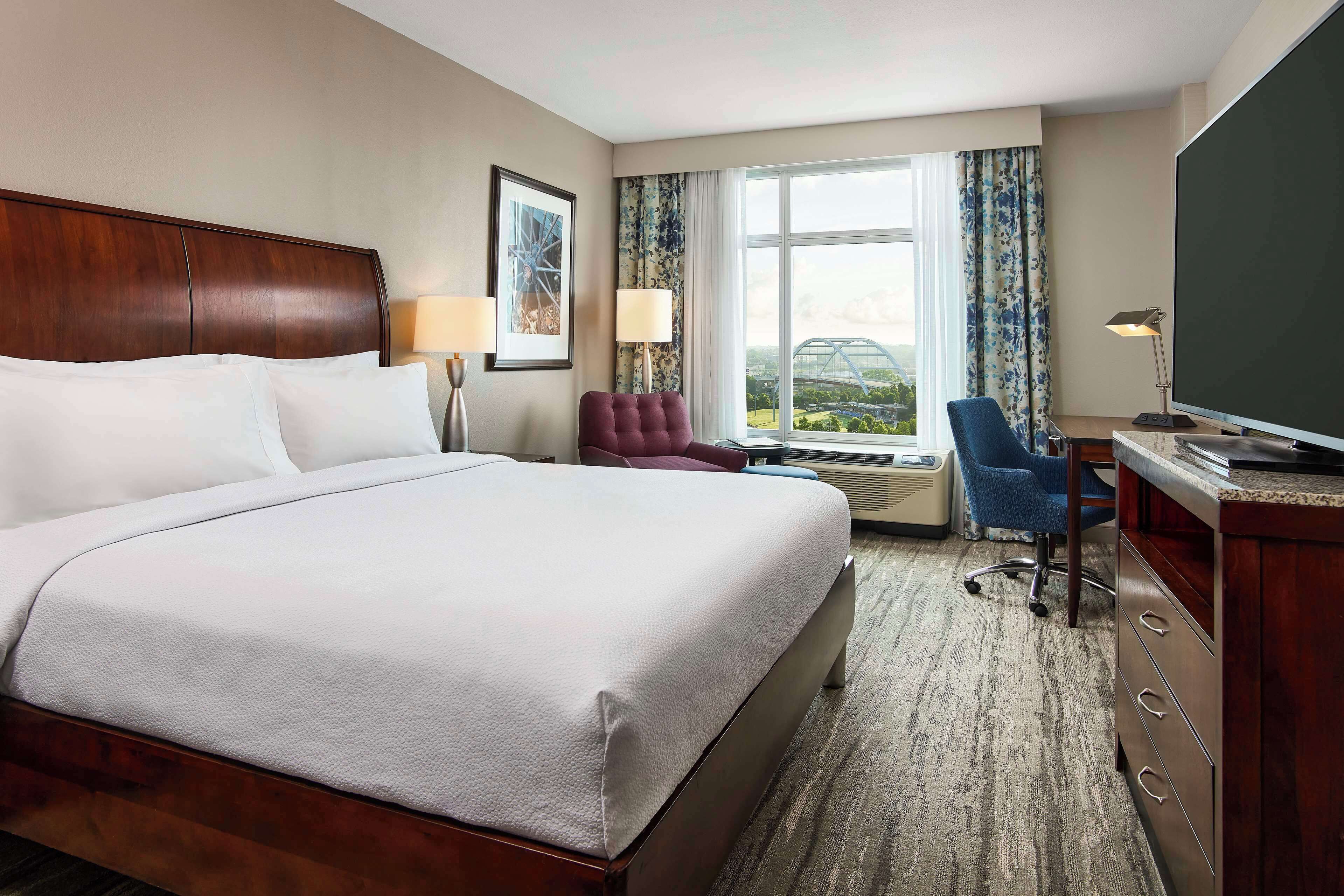 A hotel room with a large bed, a desk with a television, a chair, and a view of the outdoors.