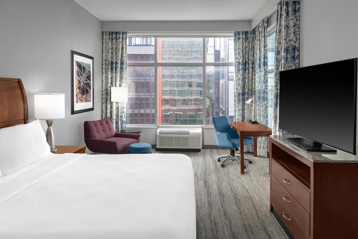 A hotel room with a bed, a desk, a chair, a TV, and a window with a city view.