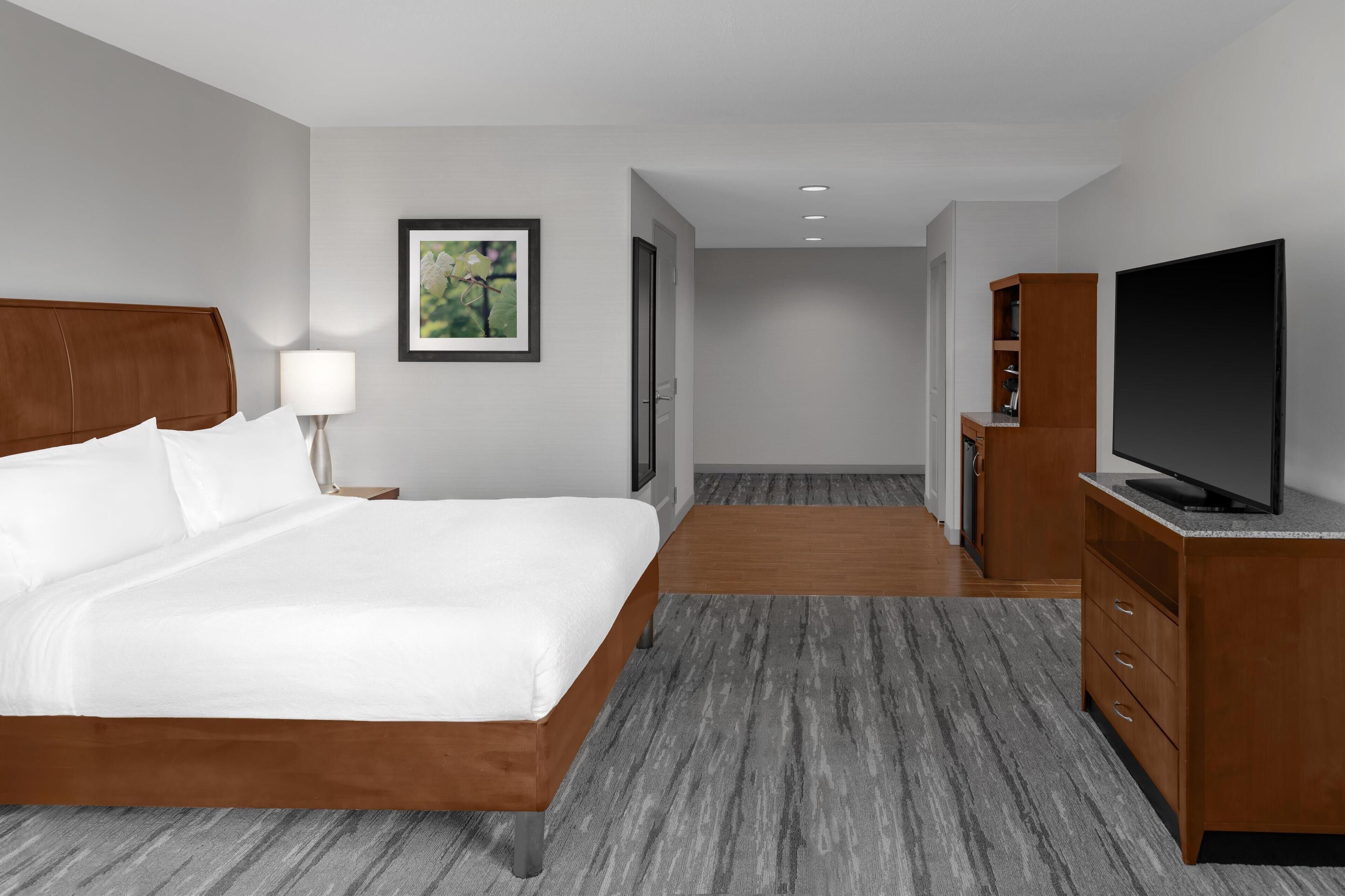A hotel room with a large bed, a TV, a wooden dresser, a lamp, and a framed picture on the wall.