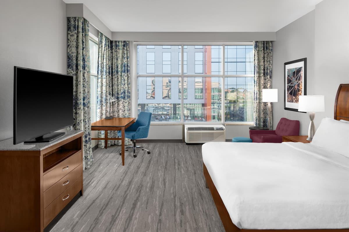A hotel room with a large bed, a desk with a chair, a flat-screen TV, a window with curtains, and a framed picture on the wall.