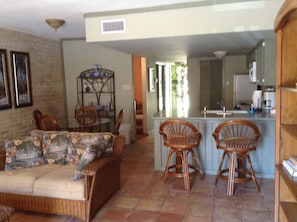 A living room with a sofa, two wooden chairs, a dining table, and a kitchen area with a sink and cabinets.
