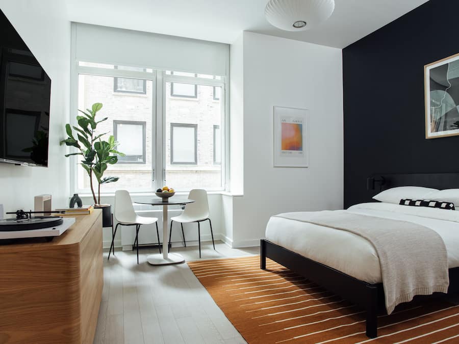 A modern bedroom with a bed, a desk with a computer, a chair, and a window with a view of buildings.