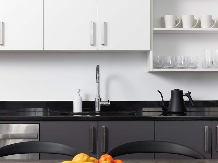 Una cocina moderna con armarios blancos, encimera negra y un lavamanos. Hay dos sillas en la mesa y un frutero con naranjas.