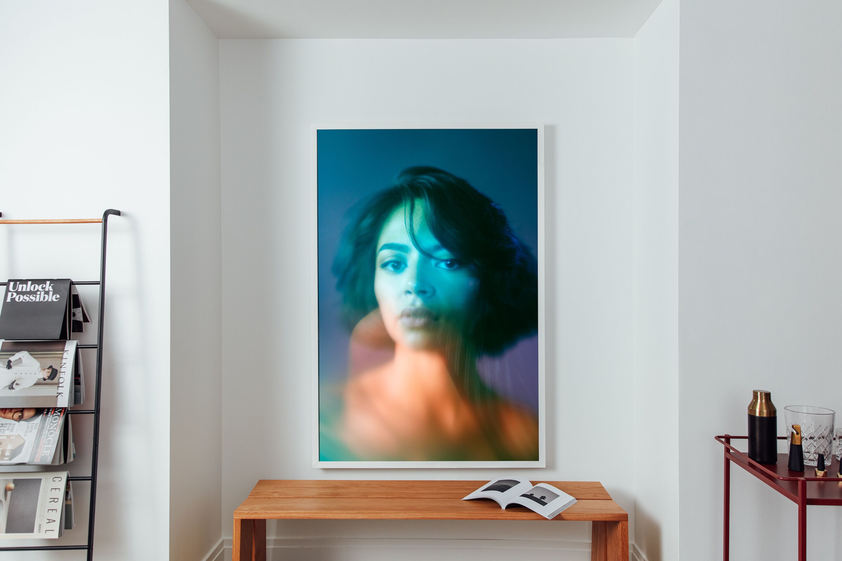 A framed portrait on a wall, a wooden side table with a book, and a metal rack with magazines.