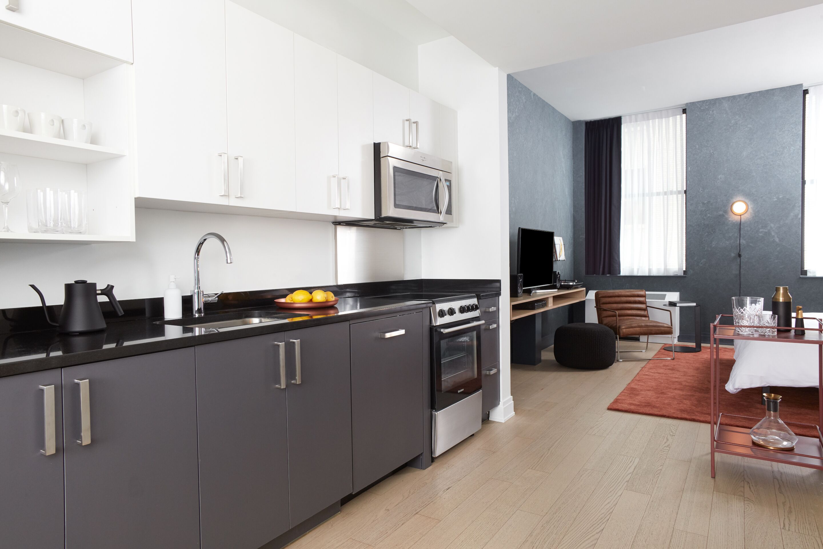 A modern kitchen with a black countertop, stainless steel appliances, and white cabinetry. There is a dining area with a table and chairs, and a large window with curtains.