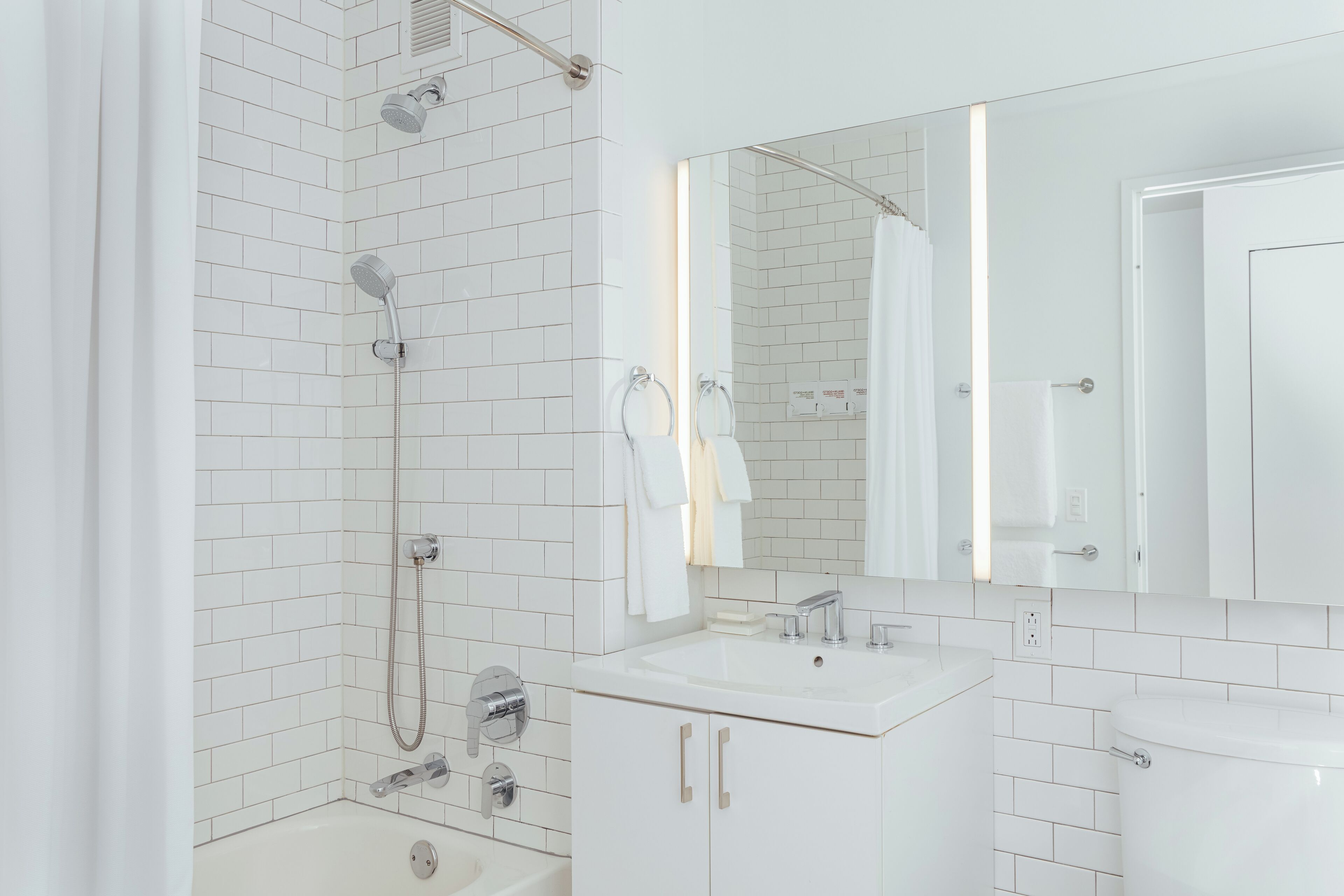 Een moderne badkamer met witte betegelde wanden, een grote spiegel, een wastafel en een douche.